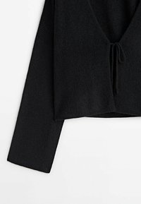 Cardigan court en tricot noir à manches longues, col en V profond et fermeture nouée à l'avant, présenté sur un fond blanc.