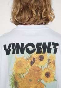 Valkoinen puuvillainen t-paita, jossa on suuri auringonkukkagrafiikka ja sana "VINCENT" lihavoidulla mustalla typografialla selän puolella.