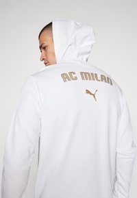 Hvid hættejakke med en kængurulomme, med guld "AC MILAN" tekst og et Puma logo på bagsiden. Blødt, glat stof og afslappet pasform.