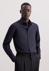 Camicetta blu navy con bottoni, maniche lunghe, colletto a punta e tessuto liscio; abbinata a pantaloni neri a pieghe.