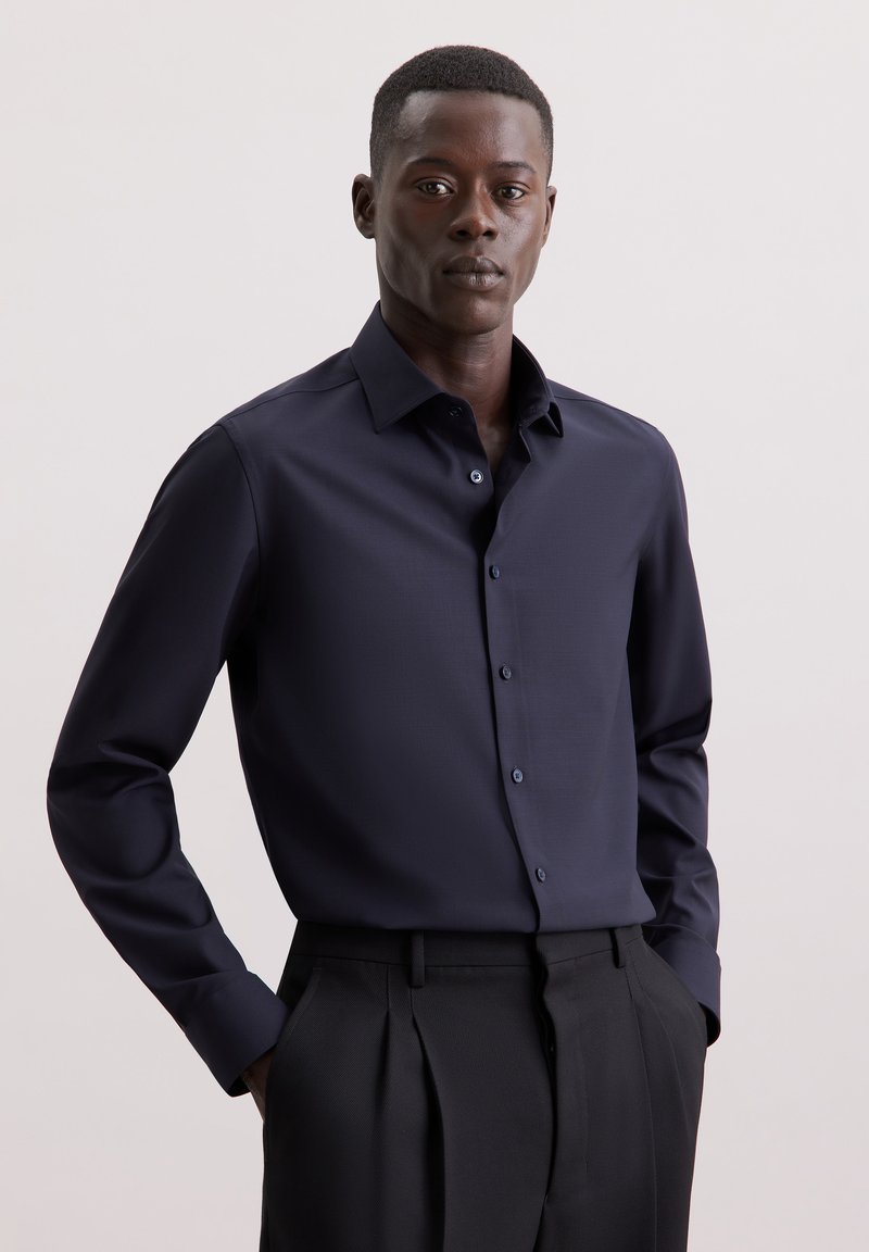 Camicetta blu navy con bottoni, maniche lunghe, colletto a punta e tessuto liscio; abbinata a pantaloni neri a pieghe.