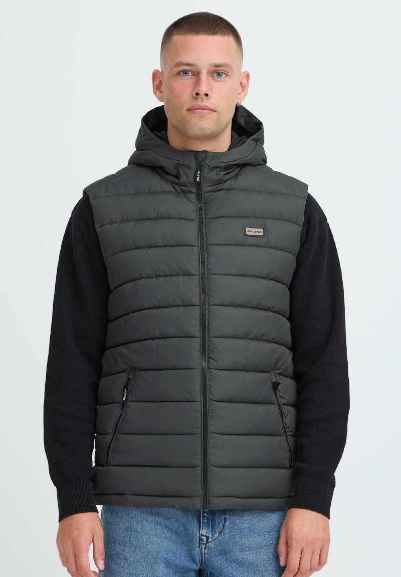 Tumehall puffer vest, millel on kapuuts, joonistatud mustri ja lukuga, külgtaskud. Ühildatud musta pika varrukaga särgiga all.