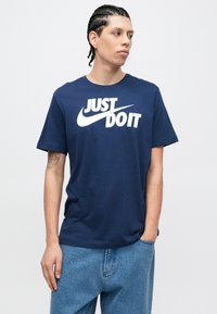 Μπλουζάκι από βαμβάκι σε σκούρο μπλε χρώμα, με μεγάλο λευκό γραφικό "Just Do It" και το λογότυπο Nike swoosh. Χαλαρή εφαρμογή με κοντά μανίκια.