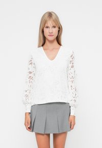 Blusa bianca in pizzo con maniche lunghe e a sbuffo, decorata con motivi floreali. Abbinata a una gonna a pieghe grigia.
