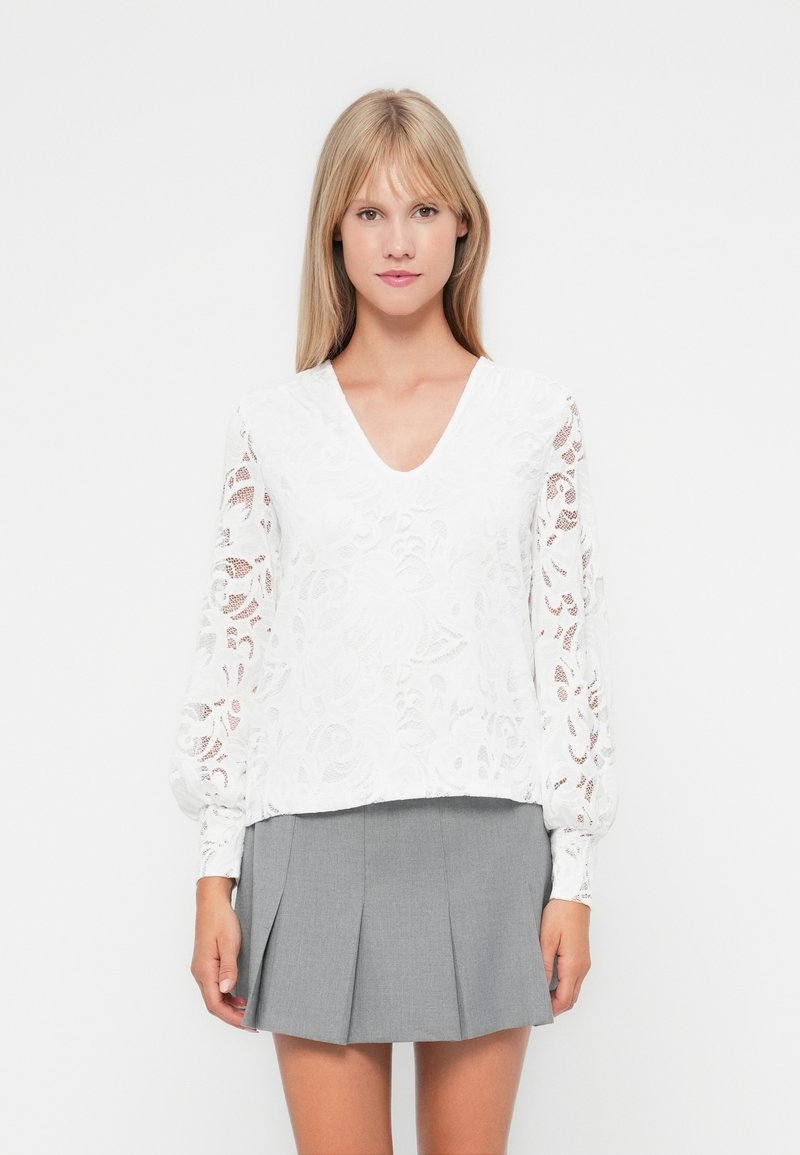 Blusa bianca in pizzo con maniche lunghe e a sbuffo, decorata con motivi floreali. Abbinata a una gonna a pieghe grigia.
