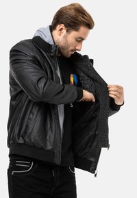 Schwarze Leder-Bomberjacke mit gestepptem Futter; grauer Fleece-Hoodie-Innenraum; gerippte Bündchen und Saum; mit Reißverschluss versehene Taschen; glatte Textur.