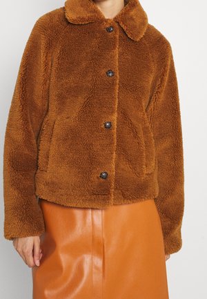 Braune, flauschige Knopfleiste mit Kragen, kombiniert mit einem hoch taillierten, glatten, orangen Lederrock, getragen von einer Person mit entspannten Händen.