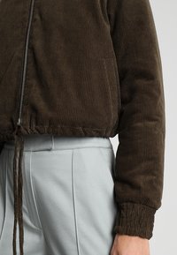 Giacca cropped in corduroy marrone con chiusura a zip e polsini elasticizzati. Presenta strisce testurizzate e un orlo con cordoncino per una vestibilità regolabile.