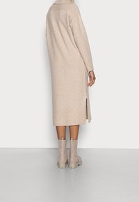 Long gilet en maille beige avec ourlet côtelé et col montant. Fente latérale. Paire de bottines beige clair avec semelles épaisses.