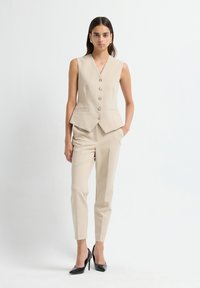 Blazer sans manches beige avec cinq boutons à l'avant, deux poches ; associé à un pantalon beige ajusté et des escarpins noirs à bout pointu. Texture lisse.