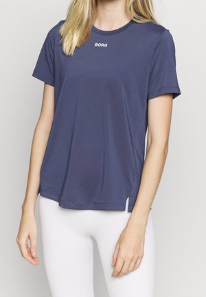 Kobieta ubrana w granatową koszulkę z krótkim rękawem z logo "BORG" na piersi oraz białe legginsy, stojąca na jasnym tle.