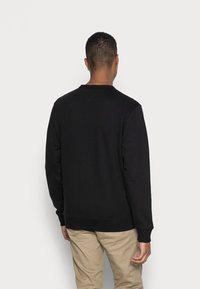 Sudadera negra de manga larga, con cuello redondo de canalé y puños. Diseño liso, sin patrones visibles. Combinada con pantalones de un beige claro.