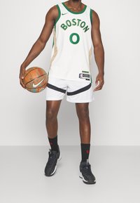 Maglia da basket bianca con dettagli verdi e testo "BOSTON", abbinata a pantaloni corti bianchi con strisce nere, scarpe da ginnastica nere e un pallone da basket.