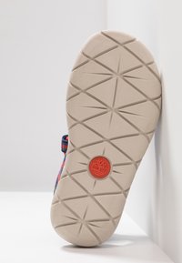 Mångfärgad sandal sula med ett mönstrat däckdesign. Har en röd accentlogo i mitten. Materialet verkar gummiliknande och hållbart.