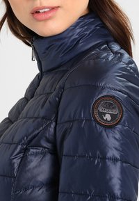 Mörkblå lättvikts puffjacka med quiltat mönster, hög krage och en cirkulär svart lapp på övre ärmen. Slät textur.