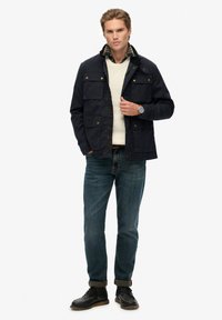 Marine waterdichte jas met koperen knopen, vier voorzakken en een geruite voering, gecombineerd met een crèmekleurige trui en blauwe jeans.