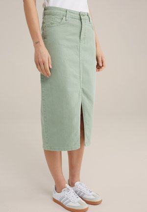 Kvinde iført en lysegrøn midi denimnederdel med frontslids, hvide sneakers og en hvid top, stående mod en ensfarvet baggrund.