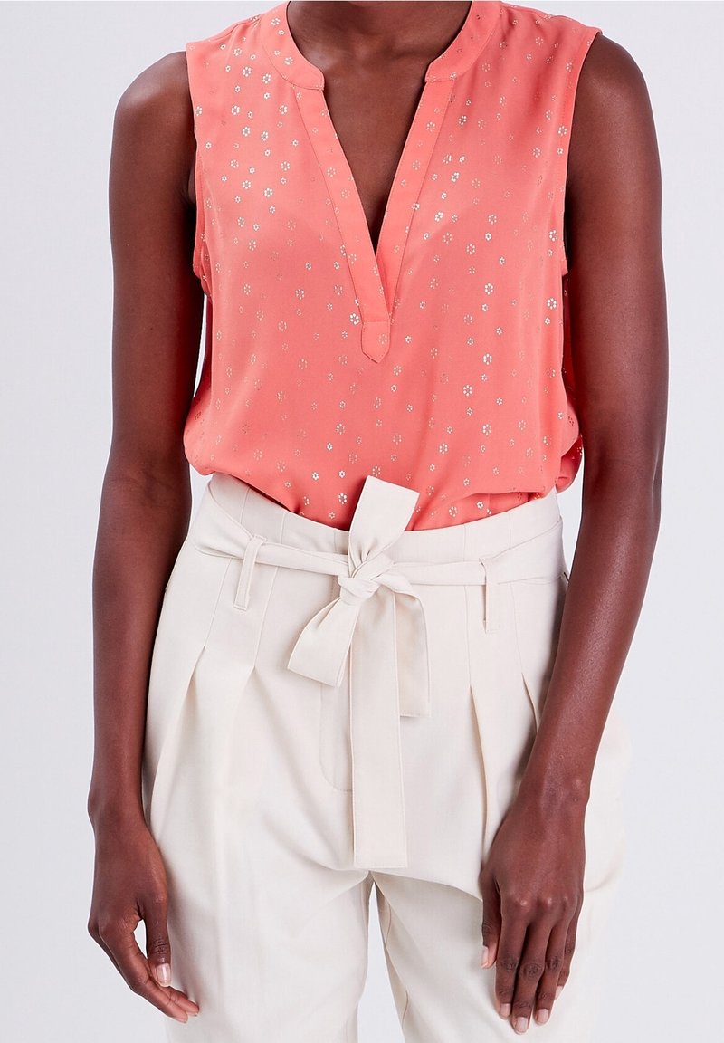 Blouse corail sans manches avec petits motifs floraux rentrée dans un pantalon beige taille haute avec une ceinture en tissu nouée.