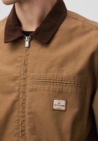 Brązowa kurtka zapinana na zamek z ciemnobrązowym kołnierzem z korduru i małą prostokątną łatą z napisem "Coalition Authentic Workwear" na kieszeni piersiowej.