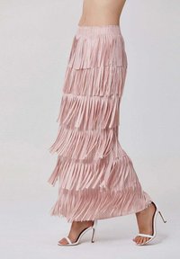 Jupe maxi frangée rose avec un design en couches, fabriquée dans un tissu doux et texturé, et dotée d'une taille ajustée. Portée avec des talons blancs à lanières.