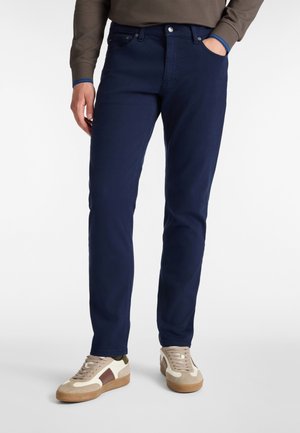 Jeans de mezclilla azul marino con corte recto. Presenta bolsillos frontales y cierre de cremallera, combinados con zapatillas beige y marrón.