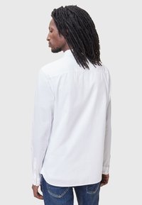 Camisa blanca de manga larga con cuello, puños abotonados y un corte relajado. Tejido suave; la parte trasera presenta detalles plizados y un dobladillo curvado.