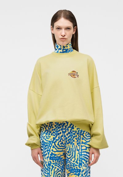 Femme portant un sweat-shirt oversize jaune pâle avec le logo « Stieglitz » et un pantalon ainsi qu'un col roulé assortis à motifs abstraits bleus et jaunes.