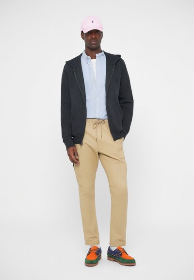 Σκοτεινό φούτερ με κουκούλα, ανοιχτό μπλε πουκάμισο με κουμπιά, μπεζ cargo παντελόνι και πολυχρωμαχα παπούτσια με ροζ καπέλο μπέιζμπολ.