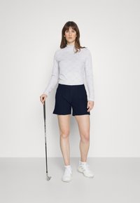 Biela dlhá rukávová blúzka s textúrovaným vzorom, kombinovaná s námorníckymi šortkami a bielymi športovými topánkami. Model drží golfovú palicu.