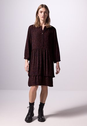Robe à manches longues, à volants, en tissu sombre avec un motif abstrait orange. Dotée d'un col et d'une ouverture à boutons sur le devant. Associée à des bottes noires lacées.
