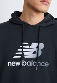 Črna kapuca iz mehke tkanine z velikim belim logotipom New Balance na prsih in ujemajočimi se vrvicami.