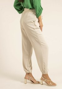 Pantalon tapered beige avec taille élastique et bas de pantalon resserrés, associé à des talons aiguilles dorés métalliques, sur un fond neutre.