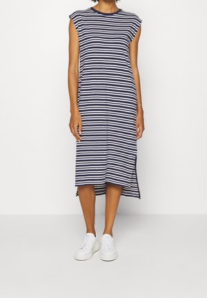 Femme portant une robe midi sans manches à rayures horizontales bleu marine et blanc, avec une fente latérale, et des baskets blanches, debout devant un fond uni.
