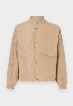 Beige tuulitakki, joka on valmistettu sileästä kankaasta. Takissa on korkea kaulus, nepparikiinnitys, kaksi läppätaskua ja elastiset hihansuut.