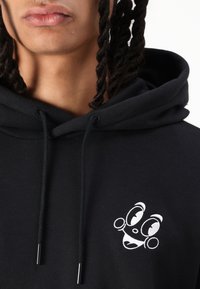 Black hooded sweatshirt featuring a small white embroidered character design on the chest, with drawstrings and a soft texture.