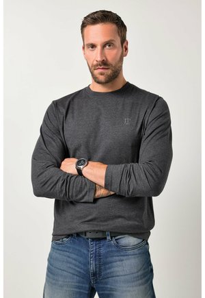 Mann mit Bart und Uhr, trägt ein dunkelgraues Langarmshirt und blaue Jeans, steht mit verschränkten Armen vor hellem Hintergrund.