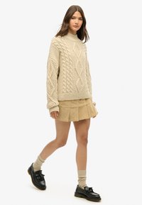 Beige chunky strikket sweater med diamantmønster, parret med en lysbrun plisseret nederdel. Sorte loafers og beige ribbede strømper fuldender looket.