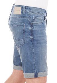 Shorts de mezclilla en azul medio con una textura ligeramente desgastada, dobladillo enrollado y detalle en el bolsillo trasero con un parche de cuero marrón.