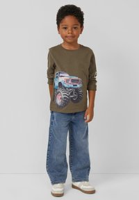 Olivgrünes Langarmshirt mit einem blauen Monstertruck-Graphic, kombiniert mit lockeren blauen Jeans und weißen Sneakers mit braunen Akzenten.
