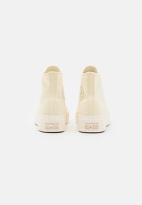 Beige korkeavartiset tennarit, jotka on valmistettu kankasta, kumipohjalla ja joissa on kohokuvioitu 'CONVERSE ALL STAR' -brändäys kantapäässä ja sivulogossa.