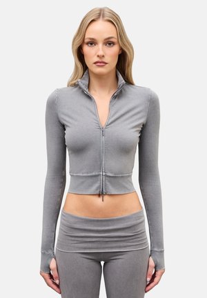 Mujer con cabello largo y rubio que lleva un top corto gris ajustado con cremallera y leggings de cintura alta a juego, de pie con los brazos relajados.