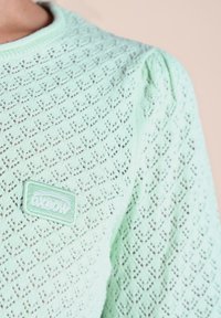 Pull en tricot vert menthe avec un motif en diamants texturé, comportant un petit patch logo Oxbow sur le devant. Col rond et poignets côtelés.