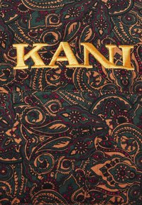 Tessuto verde e oro con un motivo paisley. Presenta "KANI" in bold, ricamato giallo su uno sfondo scuro e texturizzato.