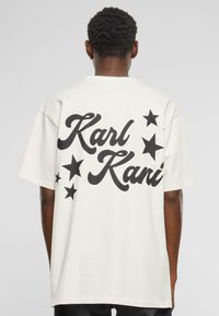 Hvit oversized t-skjorte med stor svart tekst som leser "Karl Kani" og svarte stjernemotiver på baksiden. Korte ermer, laget av bomull.