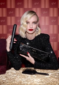 Zwarte stijltang in de ene hand, goud en zwart gestructureerde clutch in de andere hand, op een glinsterend gouden oppervlak. Model draagt een sequined zwarte top.