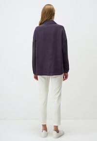 Felpa oversize viola con colletto, vestibilità comoda e maniche lunghe, abbinata a pantaloni bianchi e sneaker casual. Sfondo semplice.