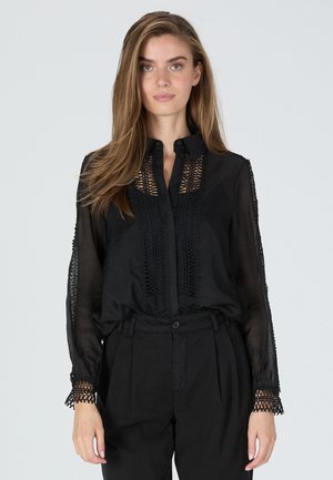 Schwarze transparente Bluse mit Spitzenverzierungen, Knopfkragen und langen Ärmeln; zeigt filigrane Muster entlang der Vorderseite und Ärmelkanten.