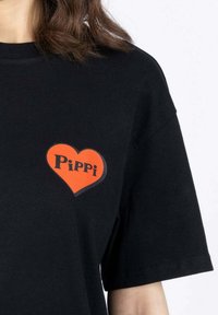Musta puuvillapaita, jossa on punainen sydänkuvio ja mustalla fontilla teksti "Pippi" vasemman rinnan alueella. Lyhyet hiharat.