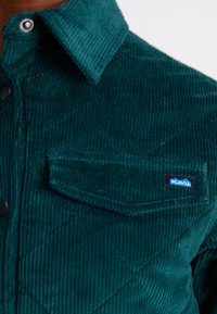 Chemise en velours côtelé turquoise avec un motif de losanges, boutons-pression, poche poitrine unique et étiquette logo KAVU. Tissu texturé avec des rayures en relief.