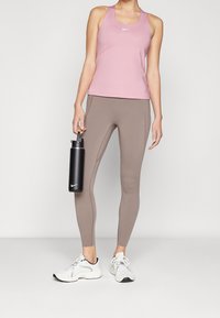 Lyserød atletisk tanktop, brune leggings og hvide sneakers. Modellen holder en sort vandflaske. Leggings har sidesømme og et tætsiddende design.